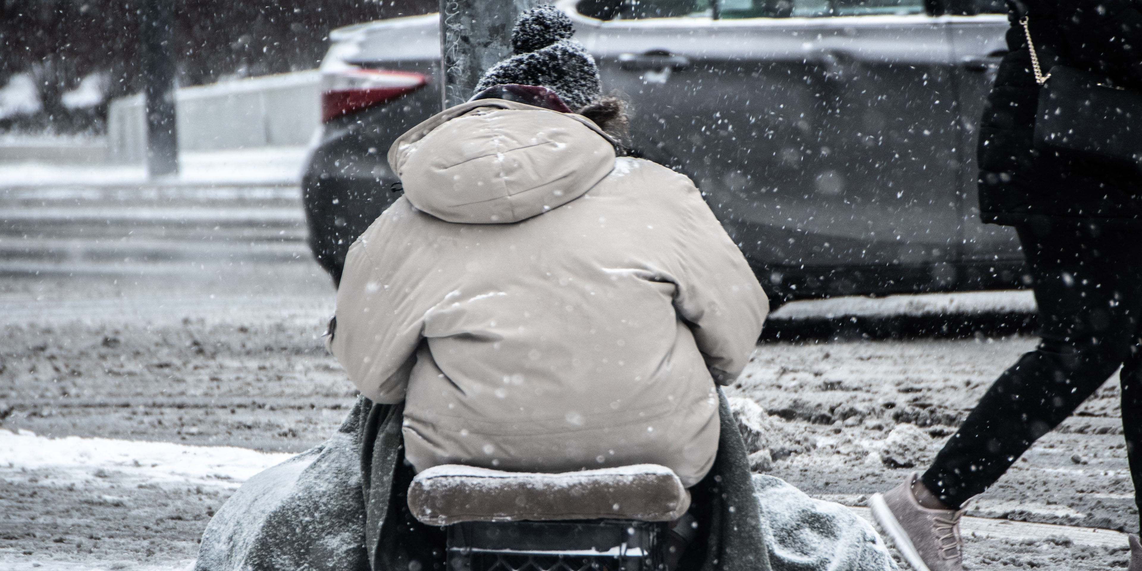 A person sitting on sidewalk during winter weather as another person walks by.